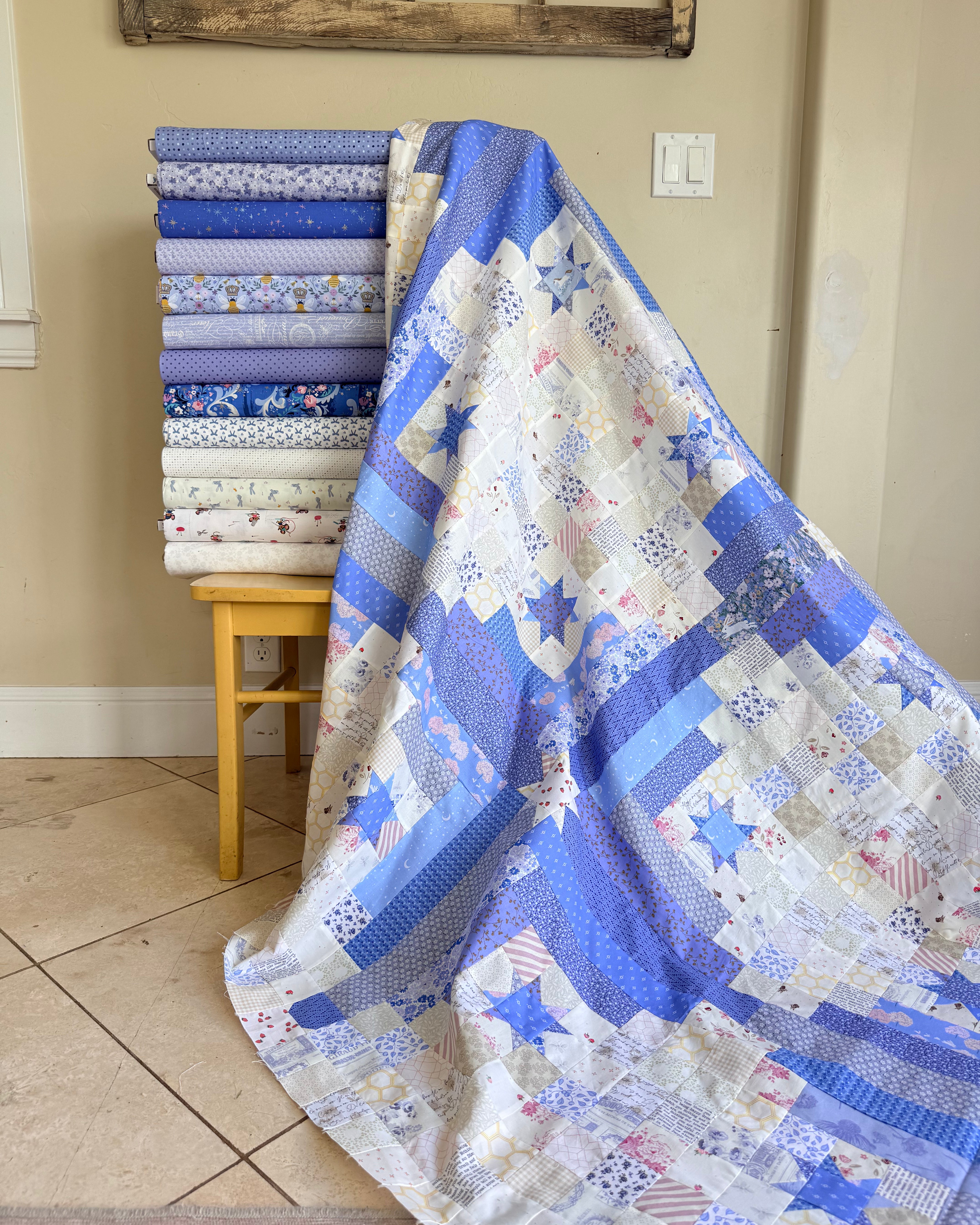 A blue and white patchwork quilt with star and geometric patterns draped over a small wooden stool, with a stack of folded fabric bolts in matching blue and white designs behind it in a home setting.