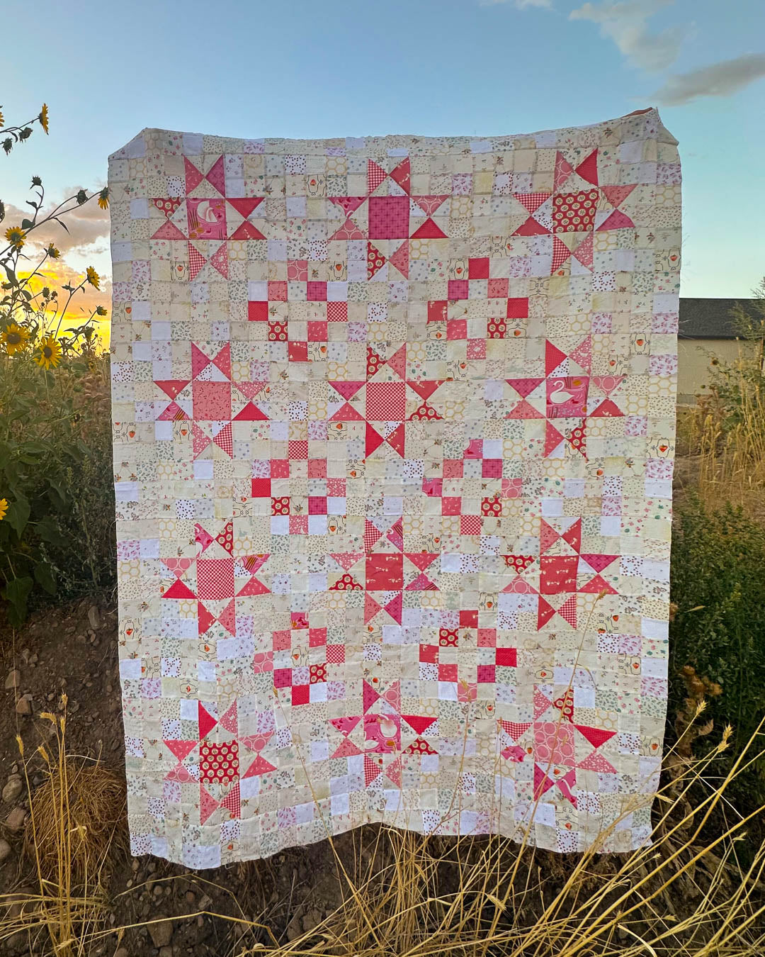 A handmade quilted blanket featuring pink and red starburst patterns on a cream-colored patchwork background, displayed outdoors in a field at sunset.