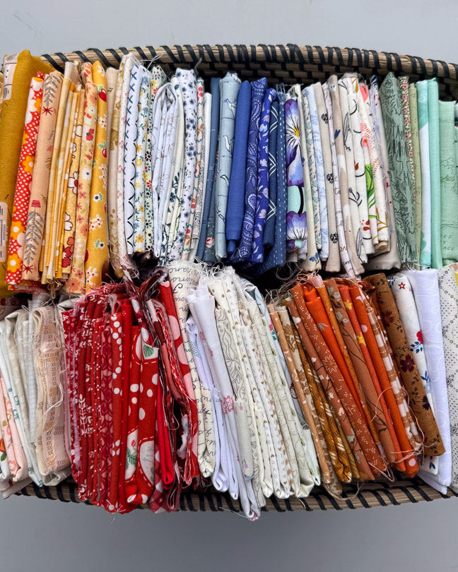 A woven basket filled with many folded fabric pieces in assorted colors and patterns, arranged in rows against a plain light gray background.