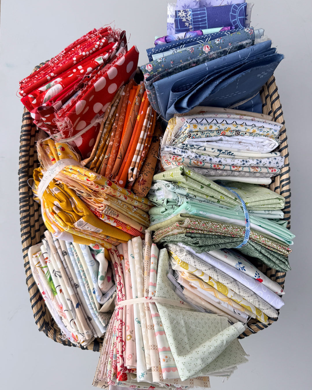 A large woven basket filled with neatly folded and bundled fabric scraps in many colors and patterns, arranged on a plain light gray background.