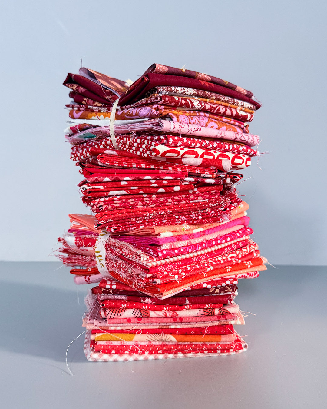 A tall stack of folded patterned fabric squares in shades of red, pink, and burgundy tied with white strings, set against a plain light gray background.