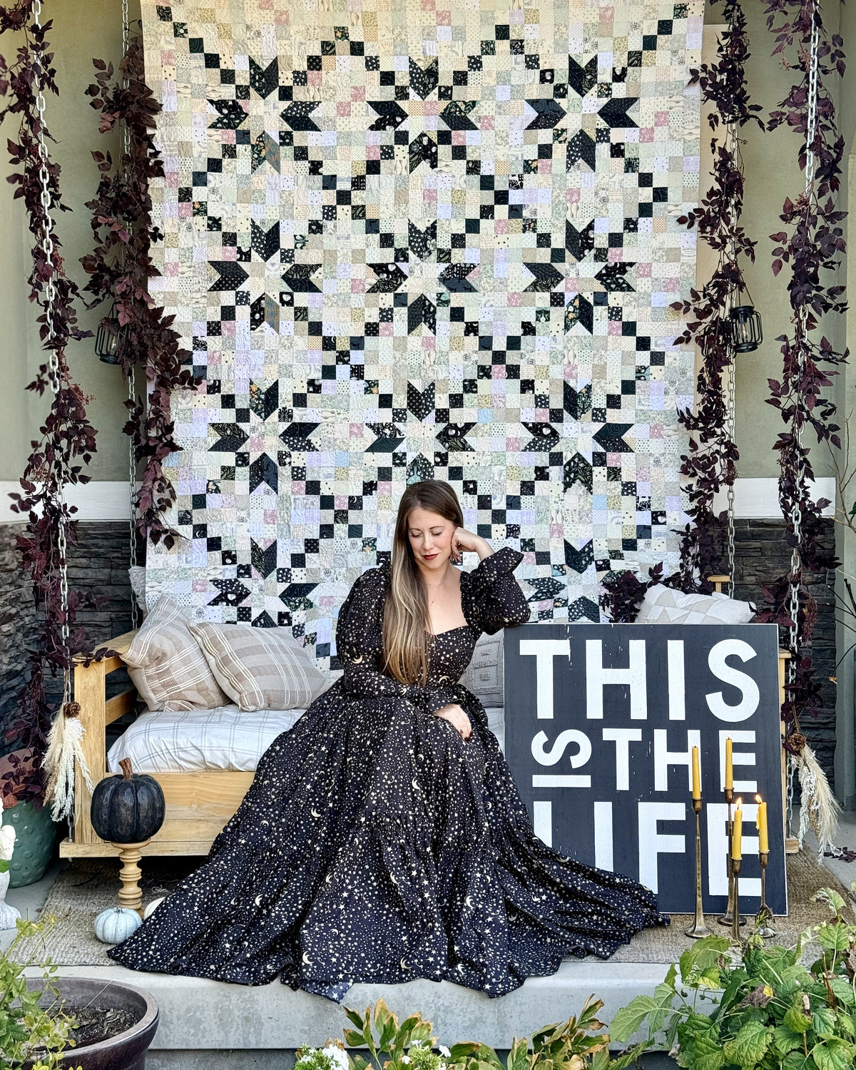 A woman in a sparkling black ballgown sits on a porch in front of a large black and cream geometric quilted wall hanging. A wooden bench with pillows is to her left, and a sign reading 'THIS IS THE LIFE' stands to her right.