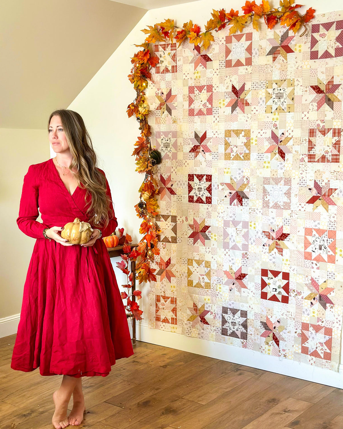 A woman in a red dress stands barefoot in a room, holding small pumpkins beside a large quilted wall hanging decorated with autumn leaves. The quilt features a patchwork design in warm red, orange, beige, and white tones.
