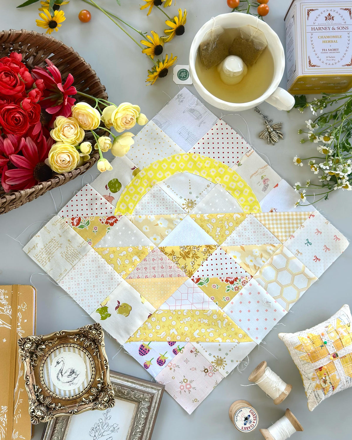 A quilted fabric square with a yellow and white geometric pattern, styled in a flat-lay arrangement with a cup of tea, flowers, spools of thread, and decorative frames on a gray surface.