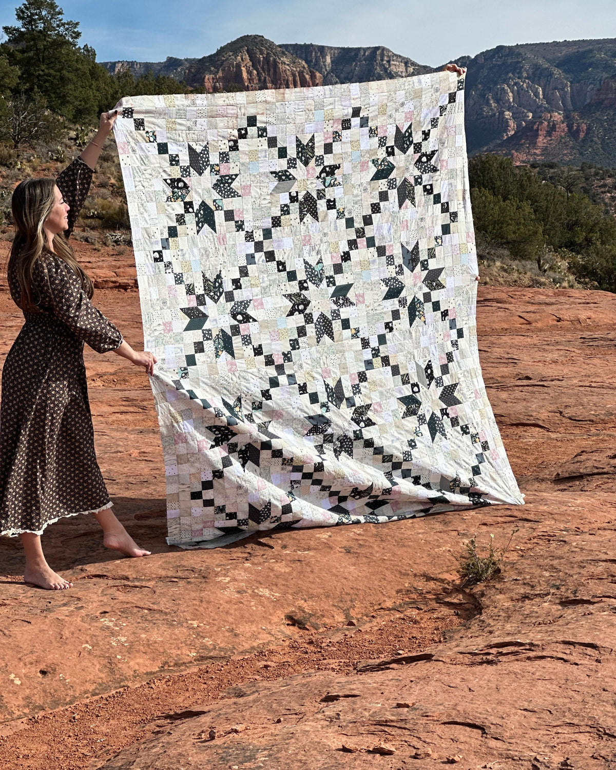 A woman in a brown polka-dot dress holds up a large quilted patchwork blanket with a black and white geometric star pattern, set against a red rock desert landscape with mountains in the background.