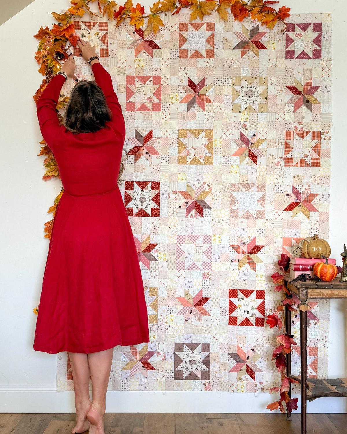 A woman in a red dress hangs autumn leaf garlands along the top edge of a large quilted wall hanging. The quilt features a patchwork design with star and geometric blocks in warm fall tones including red, orange, beige, and brown on a white background.