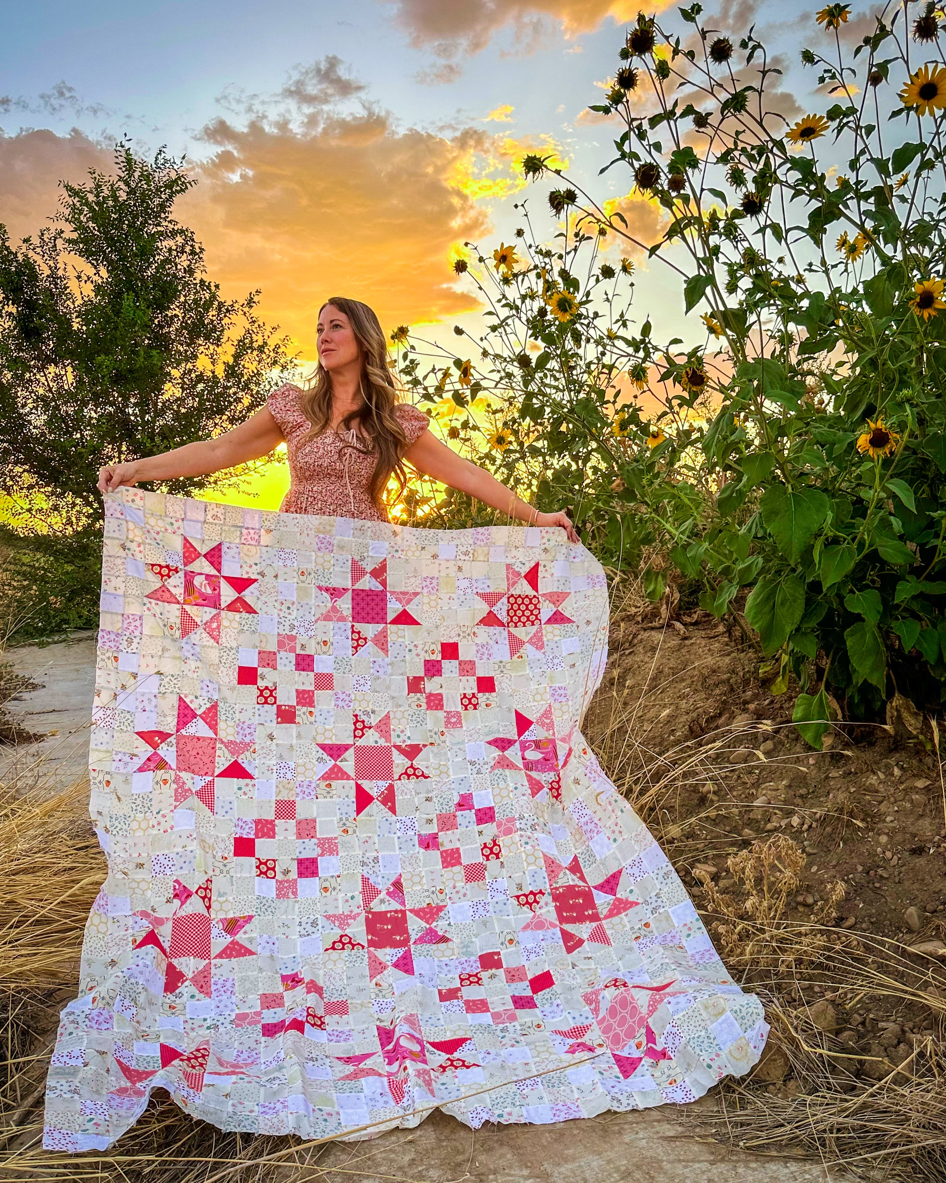A woman outdoors at sunset holds up a large quilted blanket featuring a patchwork design of pink, red, and white star and geometric blocks, with sunflowers and trees in the background.
