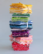 A tall stack of folded fabric bundles tied with white string, arranged vertically against a light gray studio background. The fabrics show many different colors and patterns, including yellow, green, blue, purple, pink, and red.