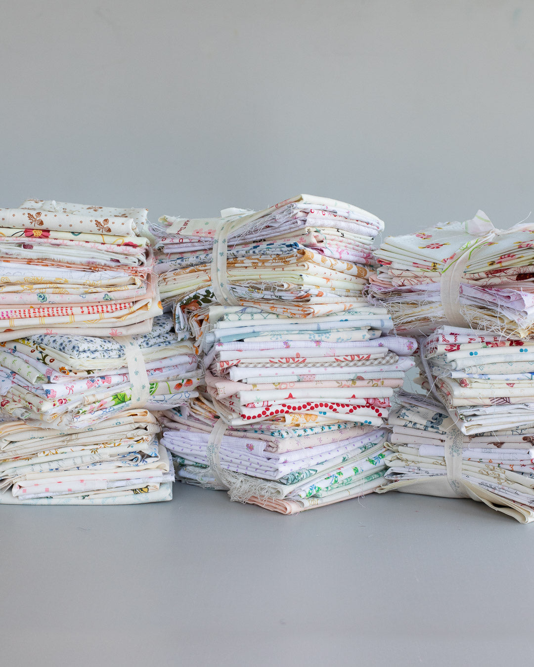 Three large stacks of folded patterned quilts or fabric bundles tied with white string, arranged on a plain light gray surface against a light gray background.
