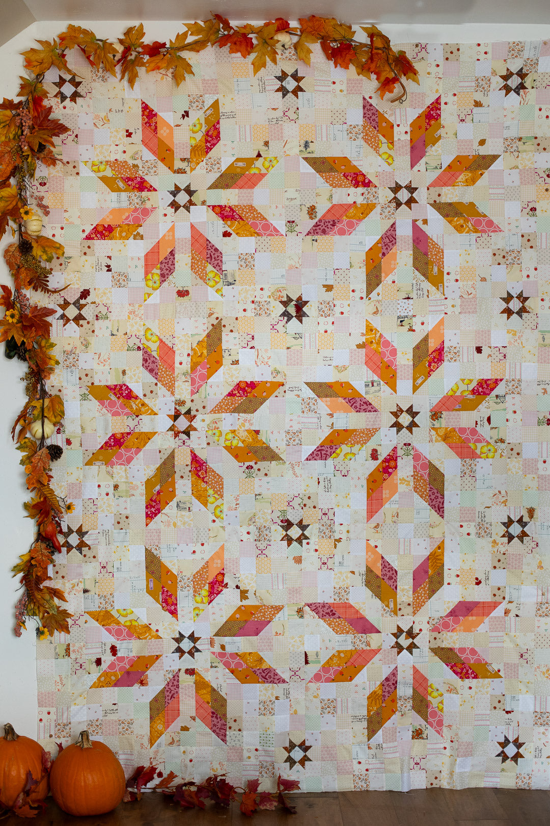 A large quilt with a repeating autumn starburst pattern in orange, pink, yellow, and brown on an off-white background. The quilt is decorated with fall leaves and a garland around the border, with two pumpkins at the bottom left.