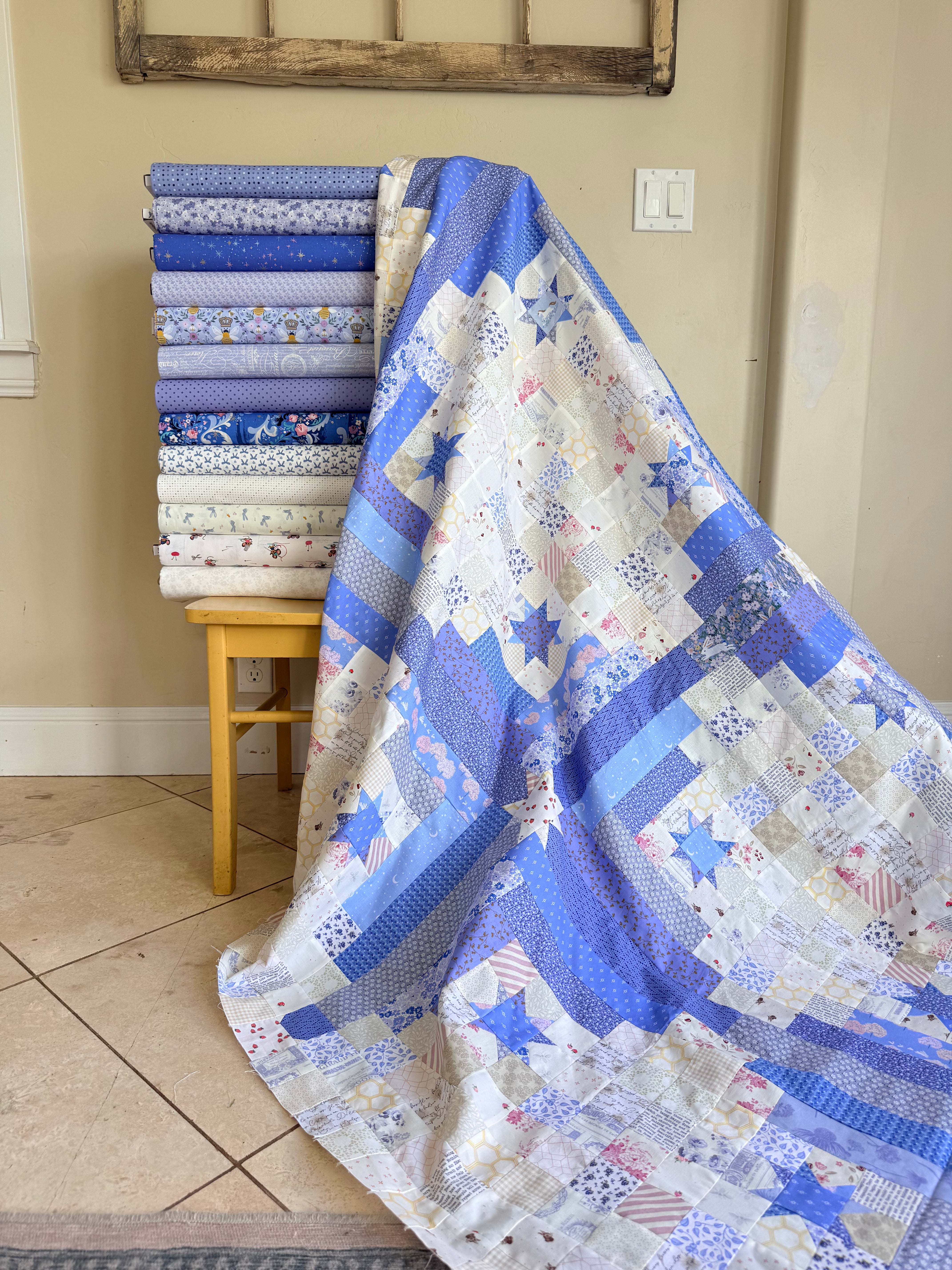A blue and white patchwork quilt with star and geometric patterns draped over a small wooden stool, with a stack of folded fabric bolts in matching blue and white designs behind it in a home setting.