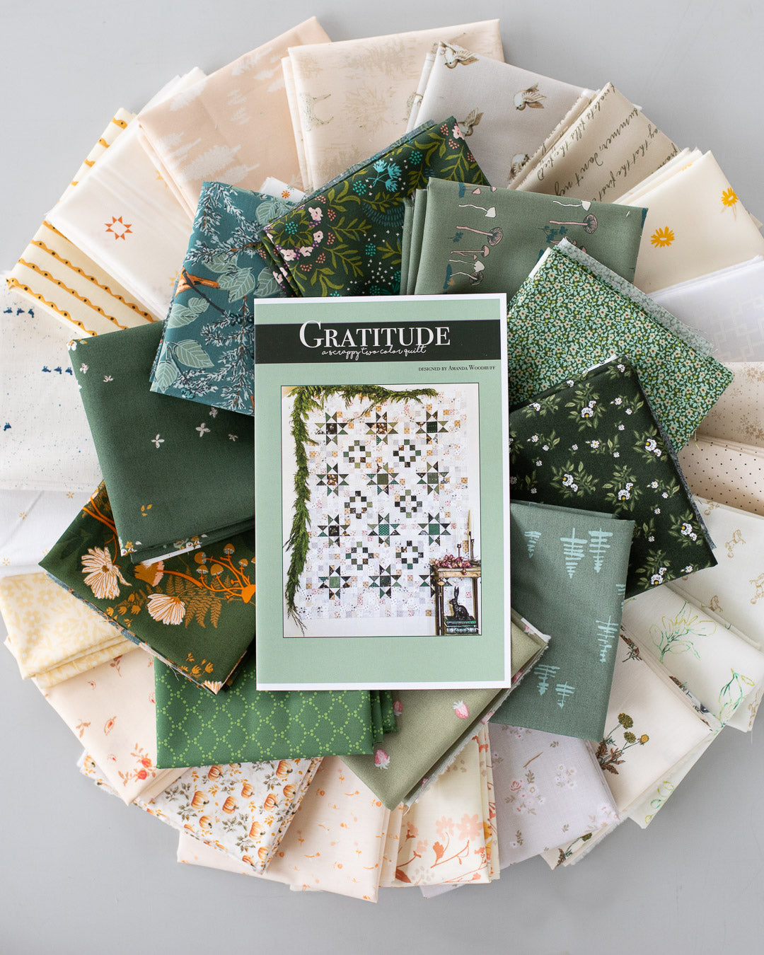 A quilt pattern book titled 'Gratitude' is centered on top of a circular arrangement of folded fabric swatches in various green, cream, and floral patterns, photographed from above on a light gray surface.