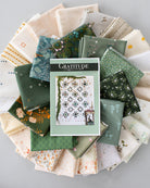 A quilt pattern book titled 'Gratitude' is centered on top of a circular arrangement of folded fabric swatches in various green, cream, and floral patterns, photographed from above on a light gray surface.
