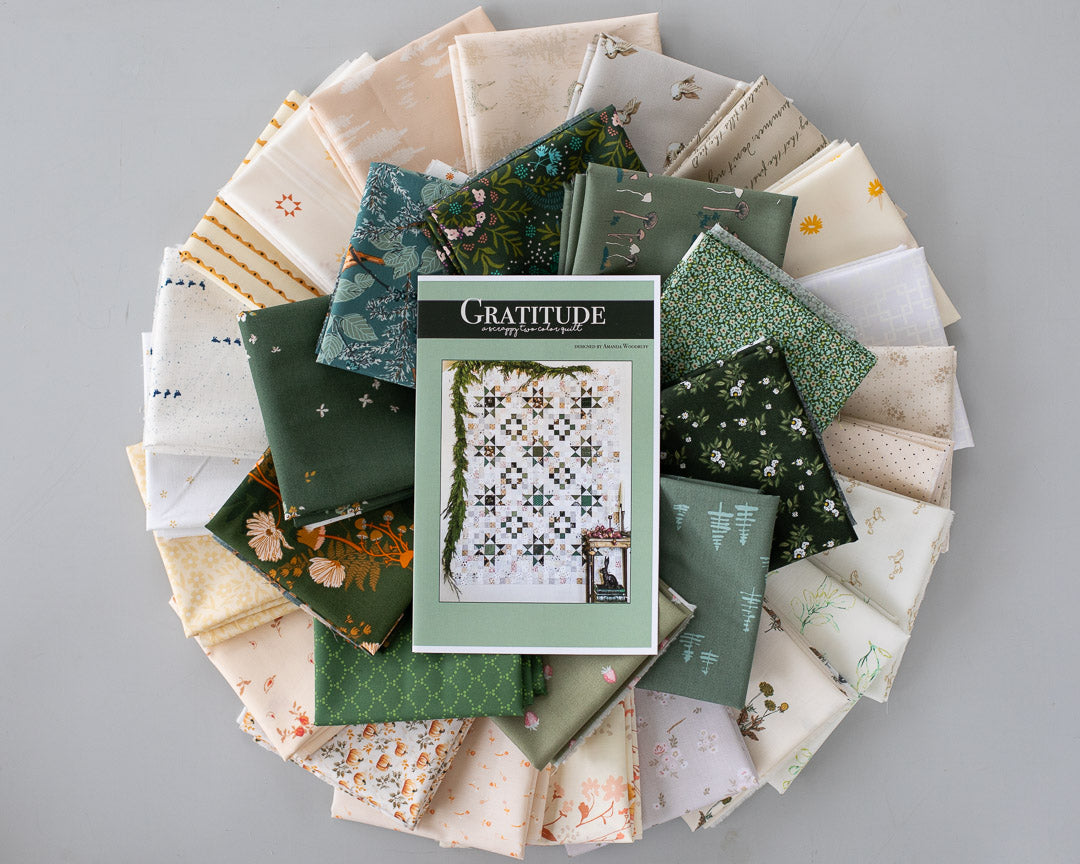 A Gratitude quilting pattern book lies on top of a circular arrangement of folded fabric swatches in various colors and patterns, including greens, creams, florals, and stripes, set against a solid gray background.