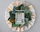 A Gratitude quilting pattern book lies on top of a circular arrangement of folded fabric swatches in various colors and patterns, including greens, creams, florals, and stripes, set against a solid gray background.