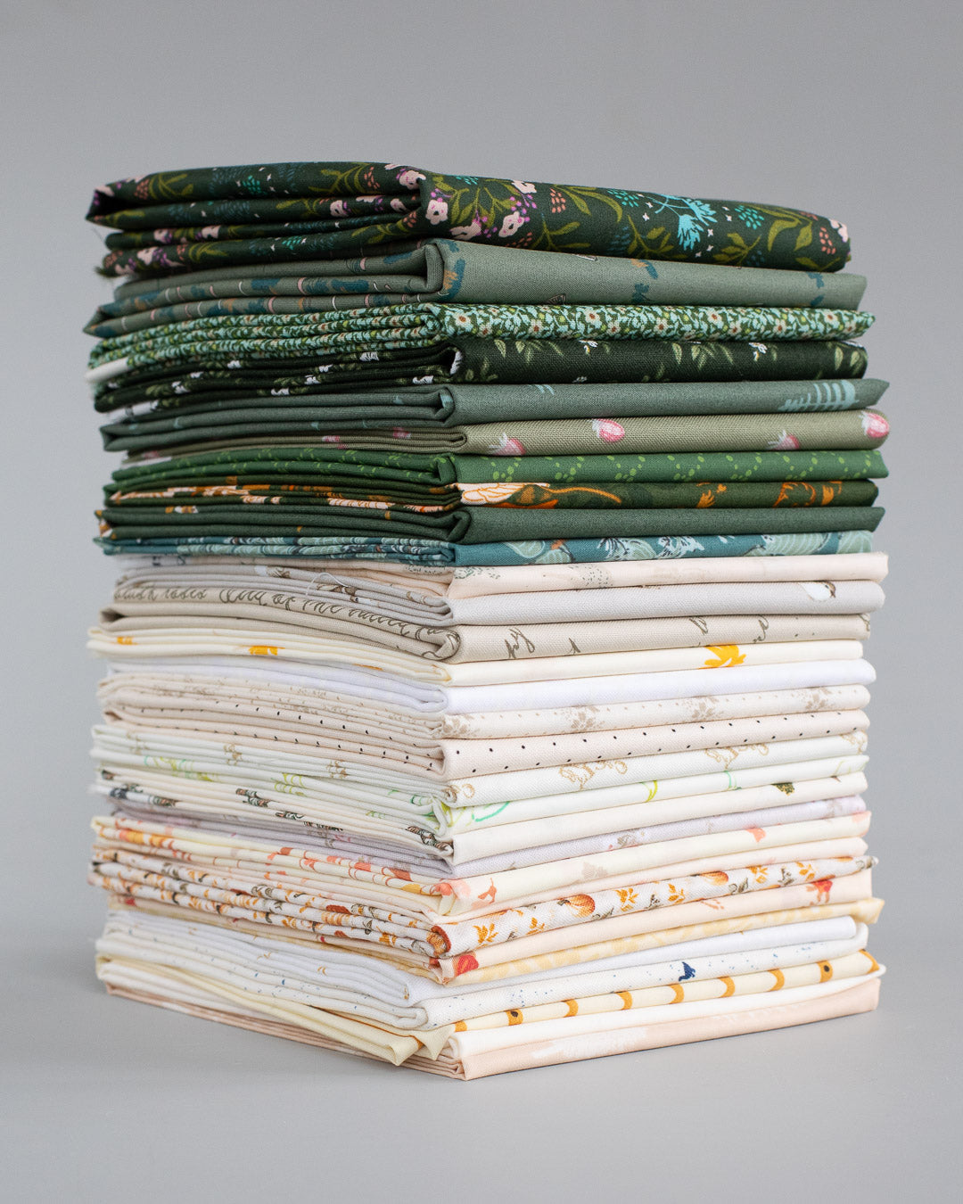 A tall stack of folded patterned fabrics in various colors and designs, including greens, creams, and florals, photographed against a neutral gray background.