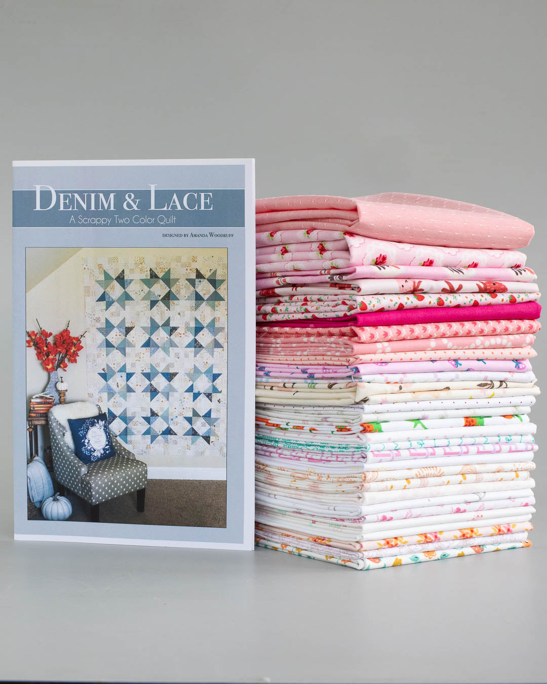 A quilt pattern book titled 'DENIM & LACE' stands next to a tall stack of folded quilts in various pink, white, and patterned designs on a neutral gray background.