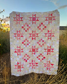 A handmade quilted blanket featuring pink and red starburst patterns on a cream-colored patchwork background, displayed outdoors in a field at sunset.