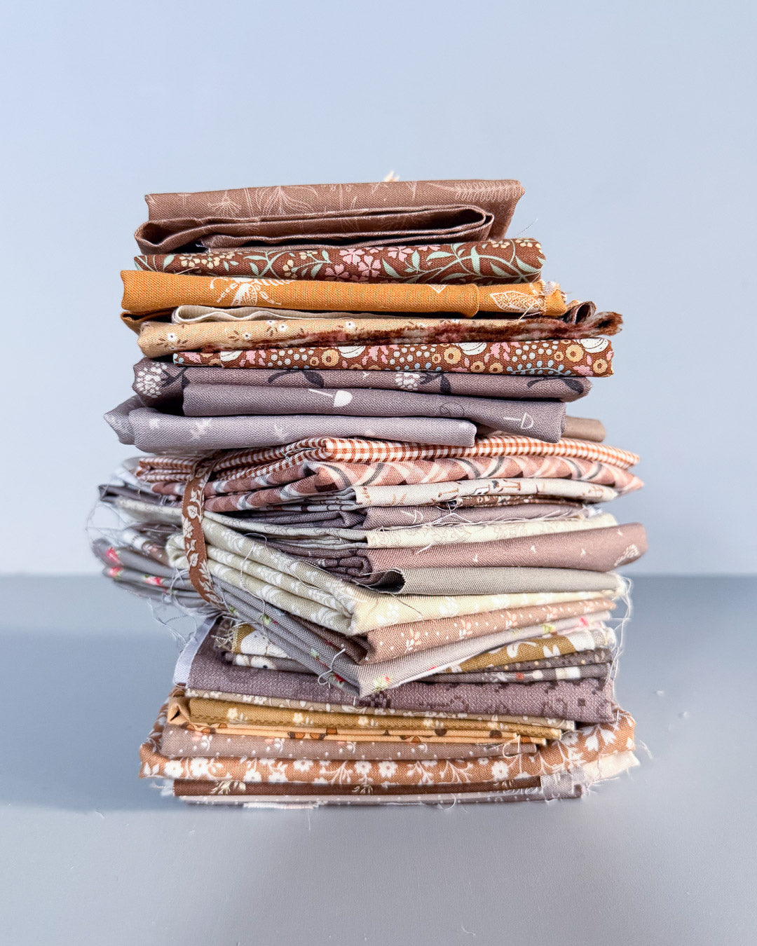 A stack of folded quilting fabrics in earthy brown, beige, and gray patterns, wrapped in clear plastic and tied with a string, set against a light gray background.