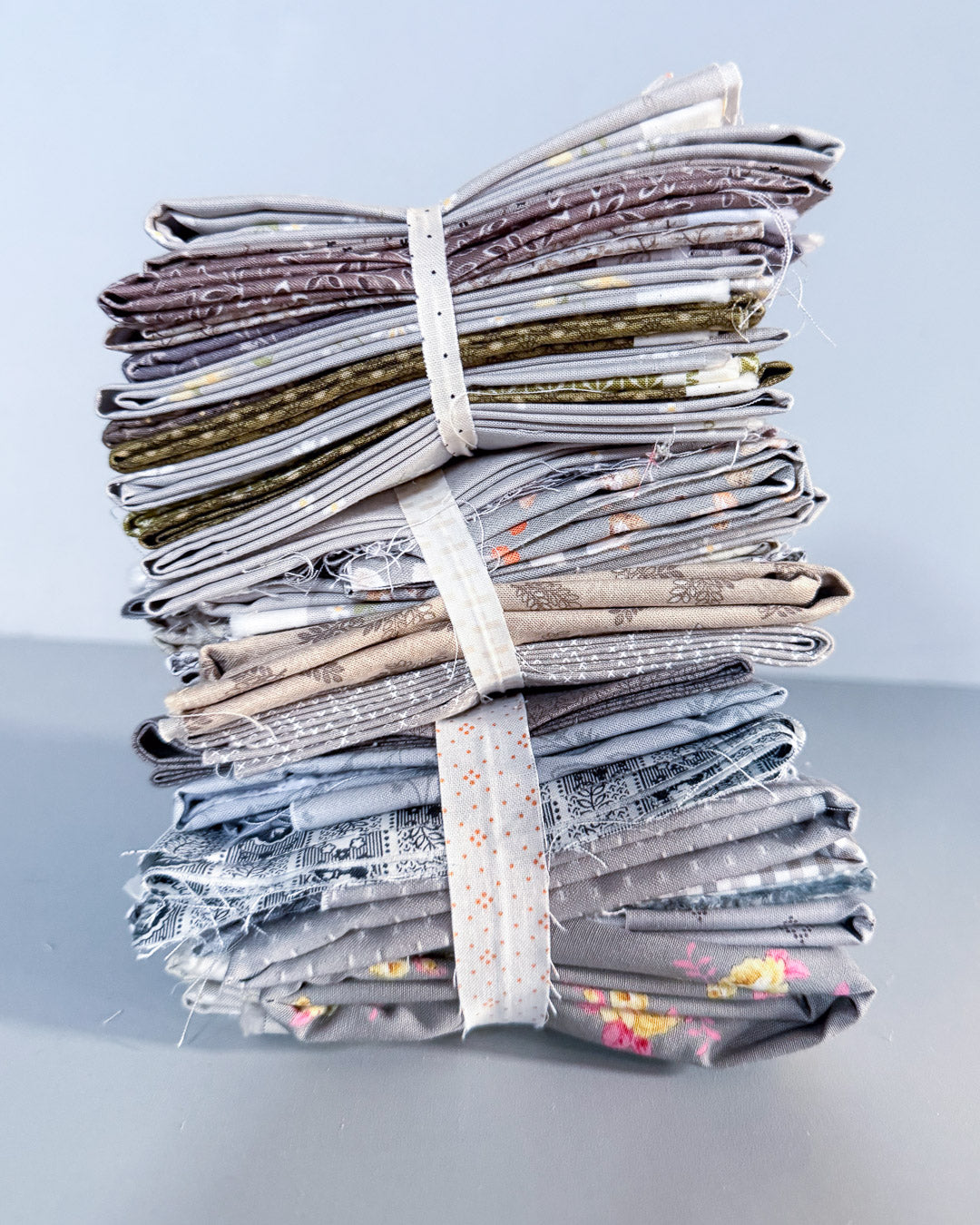 A stack of folded fabric pieces bundled together with a white elastic band, photographed on a light gray surface against a pale blue-gray background.