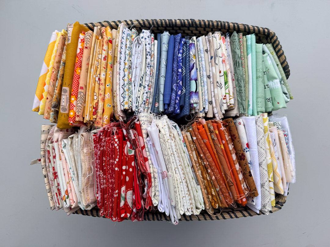 A woven basket filled with many folded fabric pieces in assorted colors and patterns, arranged in rows against a plain light gray background.