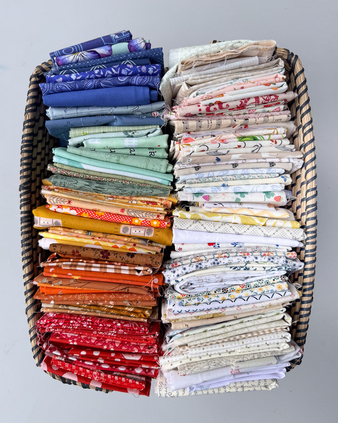 A large woven basket filled with neatly folded fabrics in a variety of colors and patterns, arranged in two columns against a plain light gray background.