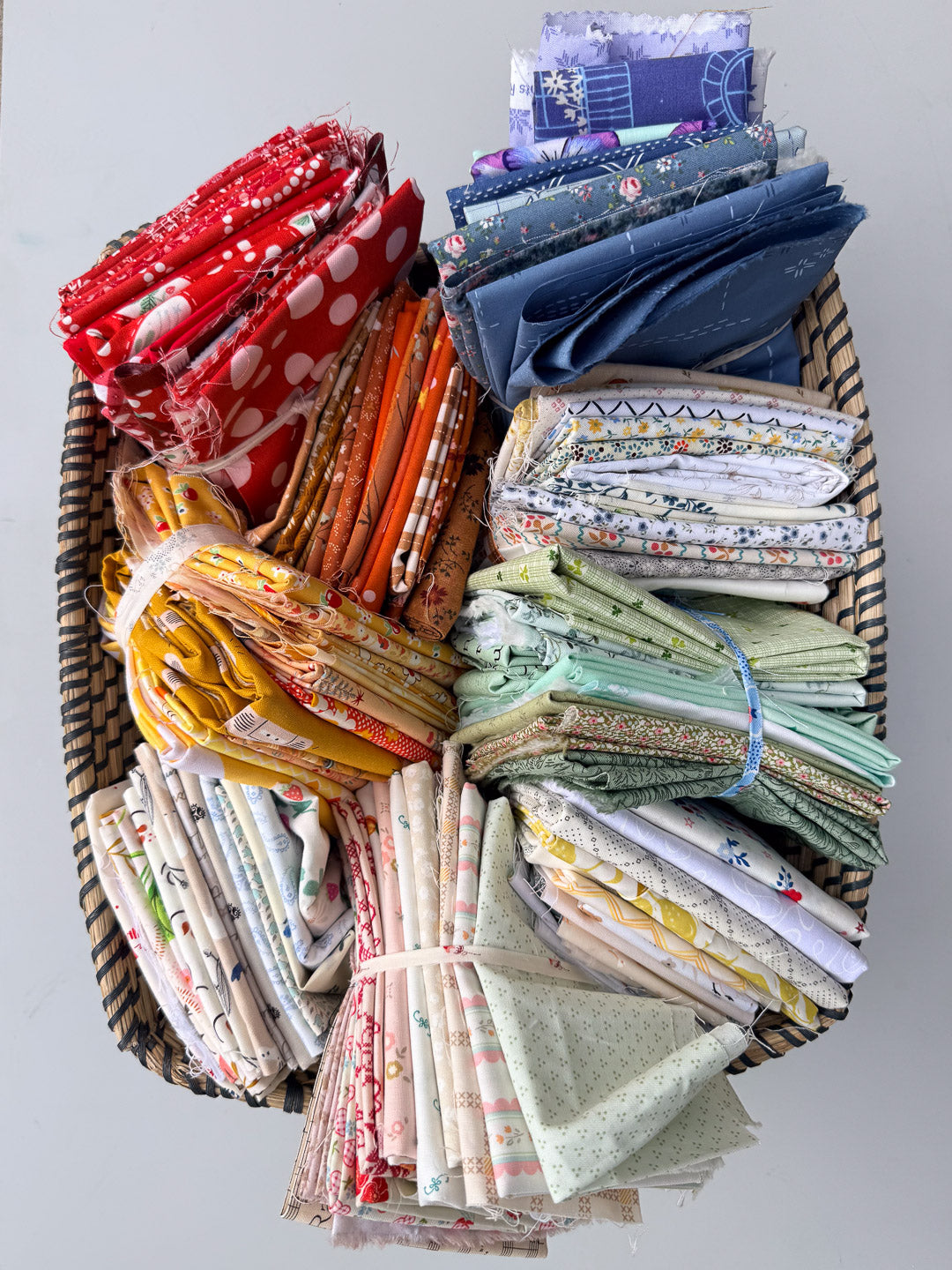 A large woven basket filled with neatly folded and bundled fabric scraps in many colors and patterns, arranged on a plain light gray background.
