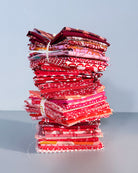 A tall stack of folded patterned fabric squares in shades of red, pink, and burgundy tied with white strings, set against a plain light gray background.
