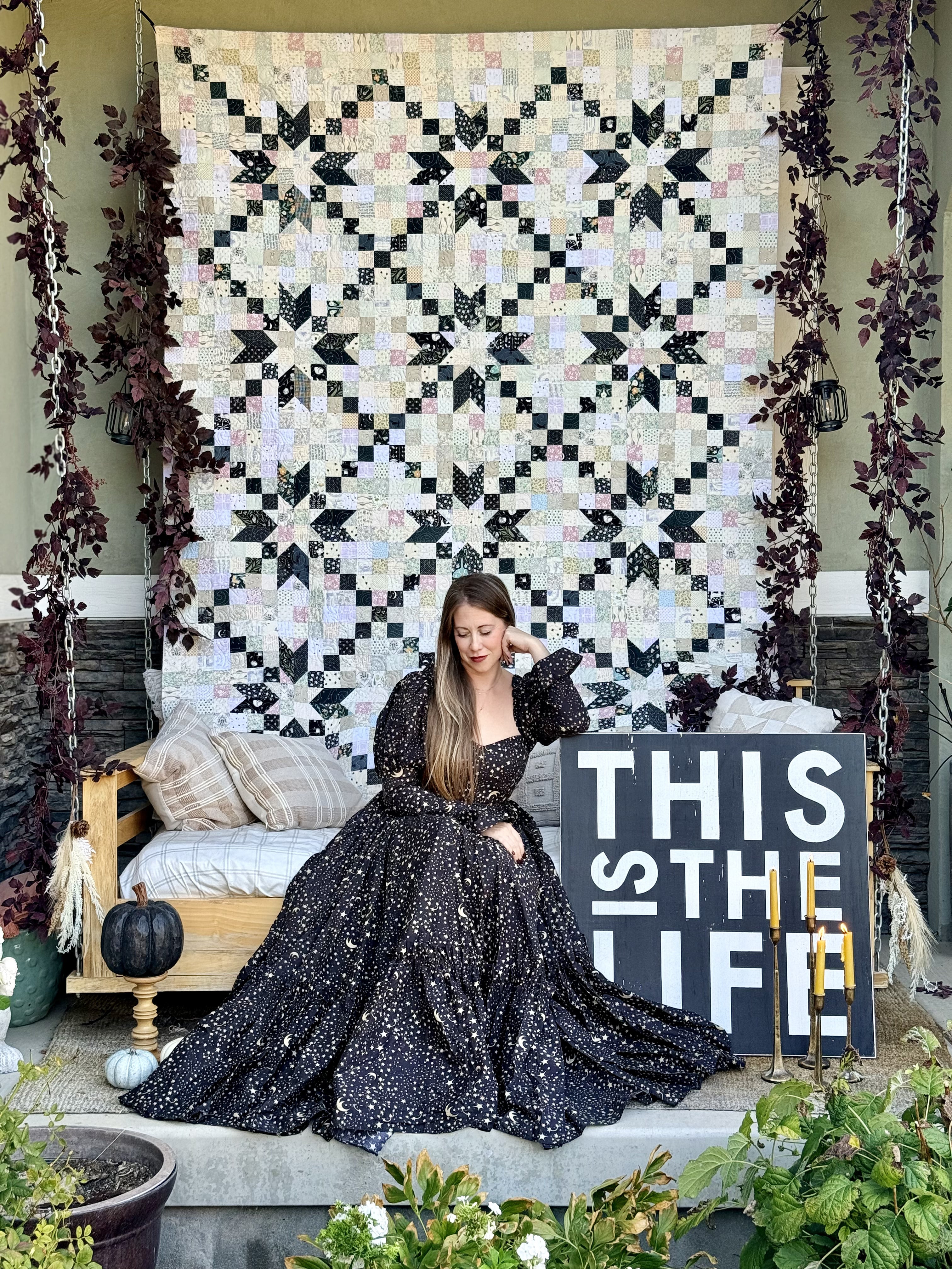 A woman in a sparkling black ballgown sits on a porch in front of a large black and cream geometric quilted wall hanging. A wooden bench with pillows is to her left, and a sign reading 'THIS IS THE LIFE' stands to her right.