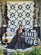 A woman in a sparkling black ballgown sits on a porch in front of a large black and cream geometric quilted wall hanging. A wooden bench with pillows is to her left, and a sign reading 'THIS IS THE LIFE' stands to her right.