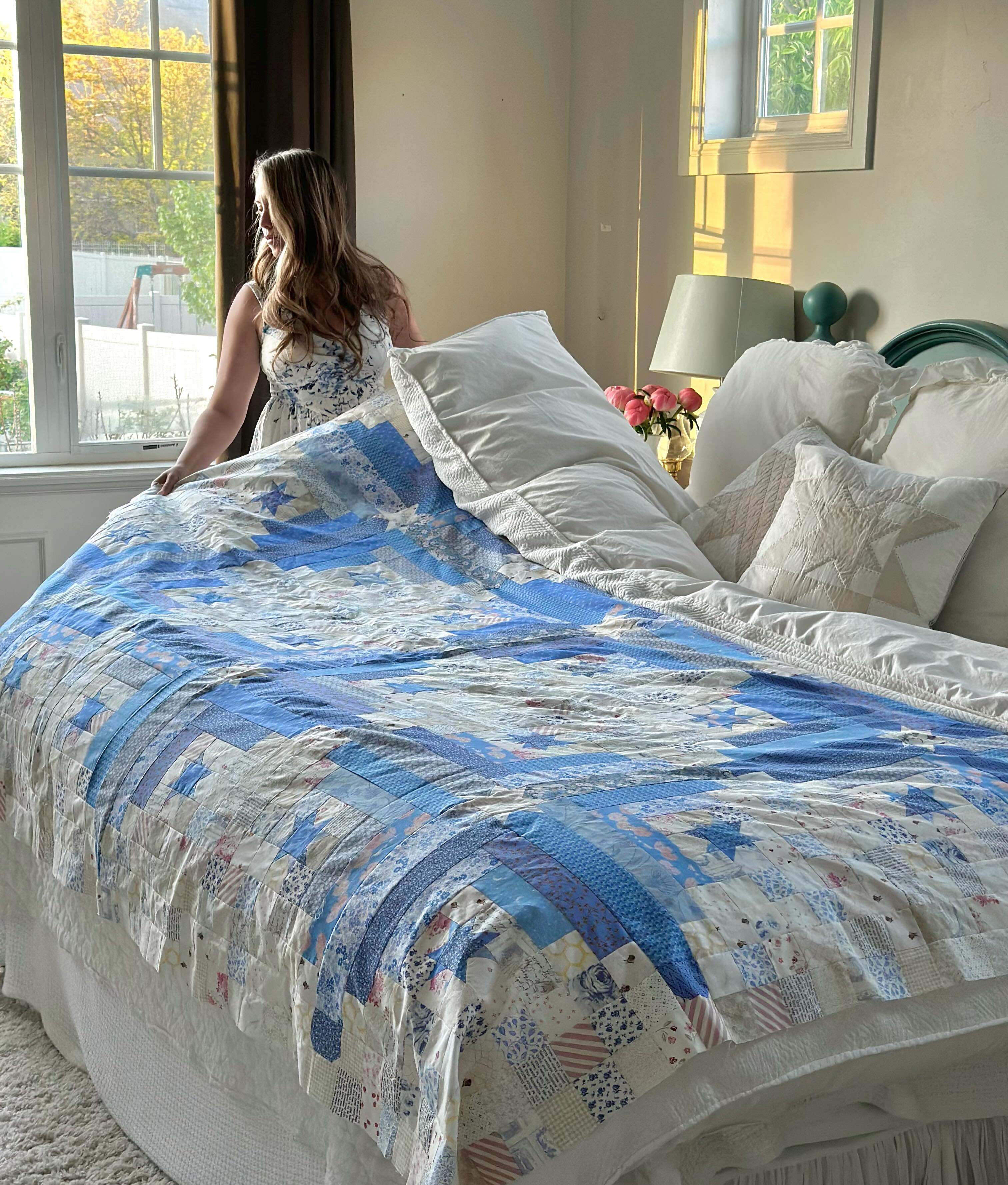 A woman in a white sleeveless dress stands at the head of a bed in a bright bedroom, holding up a patchwork quilt with blue, white, and beige sections. The bed is made with white linens and decorative pillows.