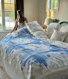 A woman in a white sleeveless dress stands at the head of a bed in a bright bedroom, holding up a patchwork quilt with blue, white, and beige sections. The bed is made with white linens and decorative pillows.
