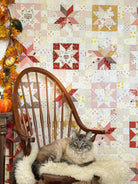 A fluffy gray and beige cat lies on a cream faux-fur throw on a wooden spindle-back chair, set against a wall covered in a patchwork quilt with autumn-themed blocks. Fall garlands and pumpkins decorate the left side of the scene.