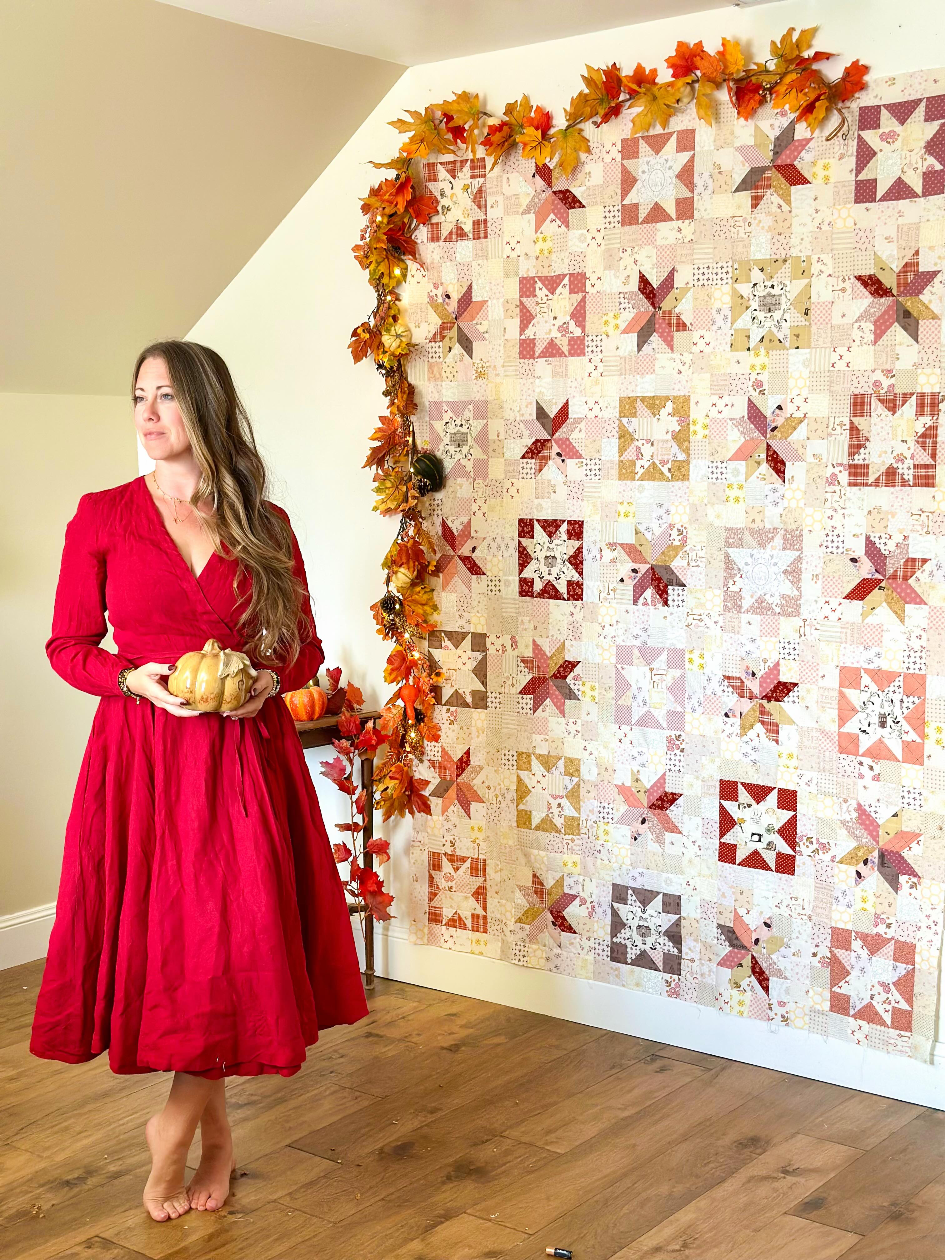 A woman in a red dress stands barefoot in a room, holding small pumpkins beside a large quilted wall hanging decorated with autumn leaves. The quilt features a patchwork design in warm red, orange, beige, and white tones.