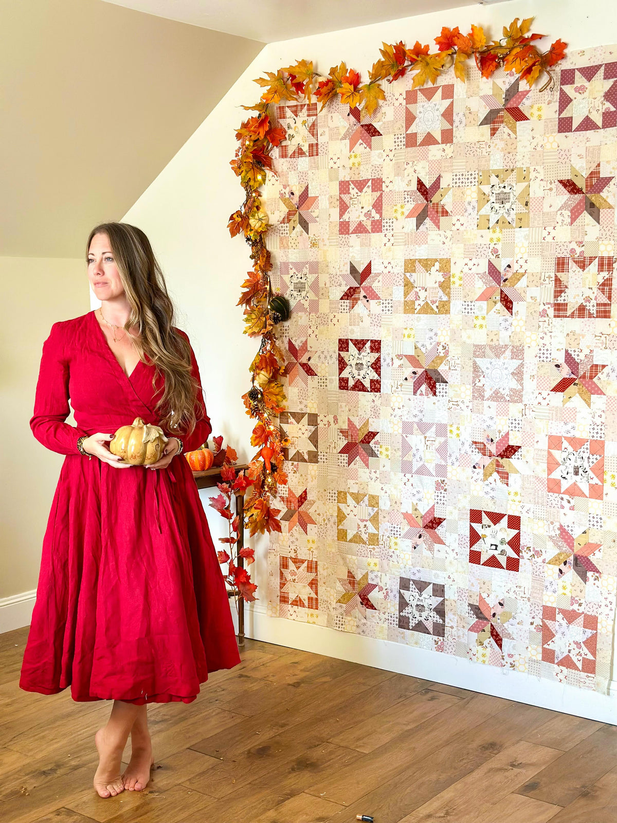 A woman in a red dress stands barefoot in a room, holding small pumpkins beside a large quilted wall hanging decorated with autumn leaves. The quilt features a patchwork design in warm red, orange, beige, and white tones.