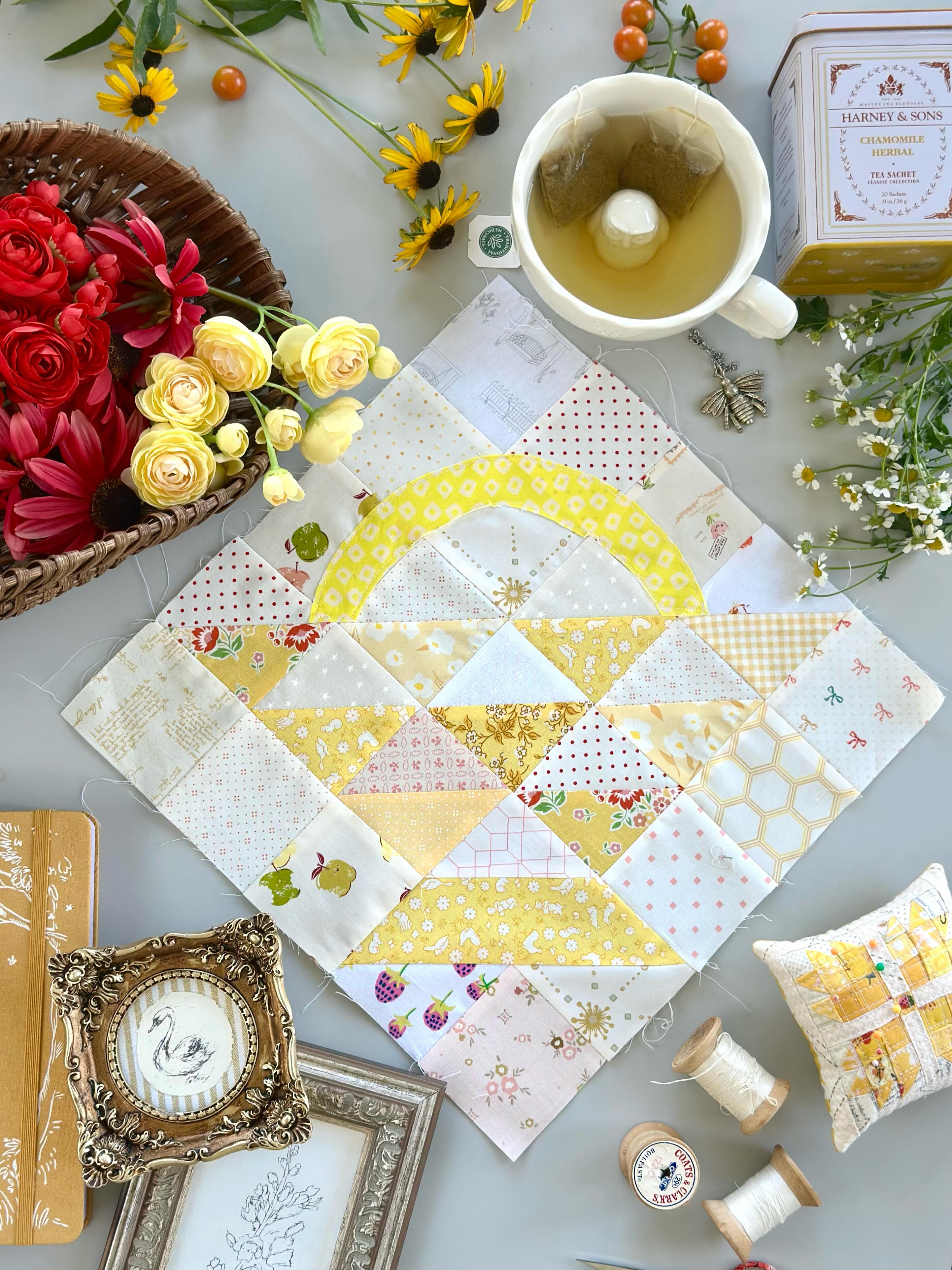 A quilted fabric square with a yellow and white geometric pattern, styled in a flat-lay arrangement with a cup of tea, flowers, spools of thread, and decorative frames on a gray surface.