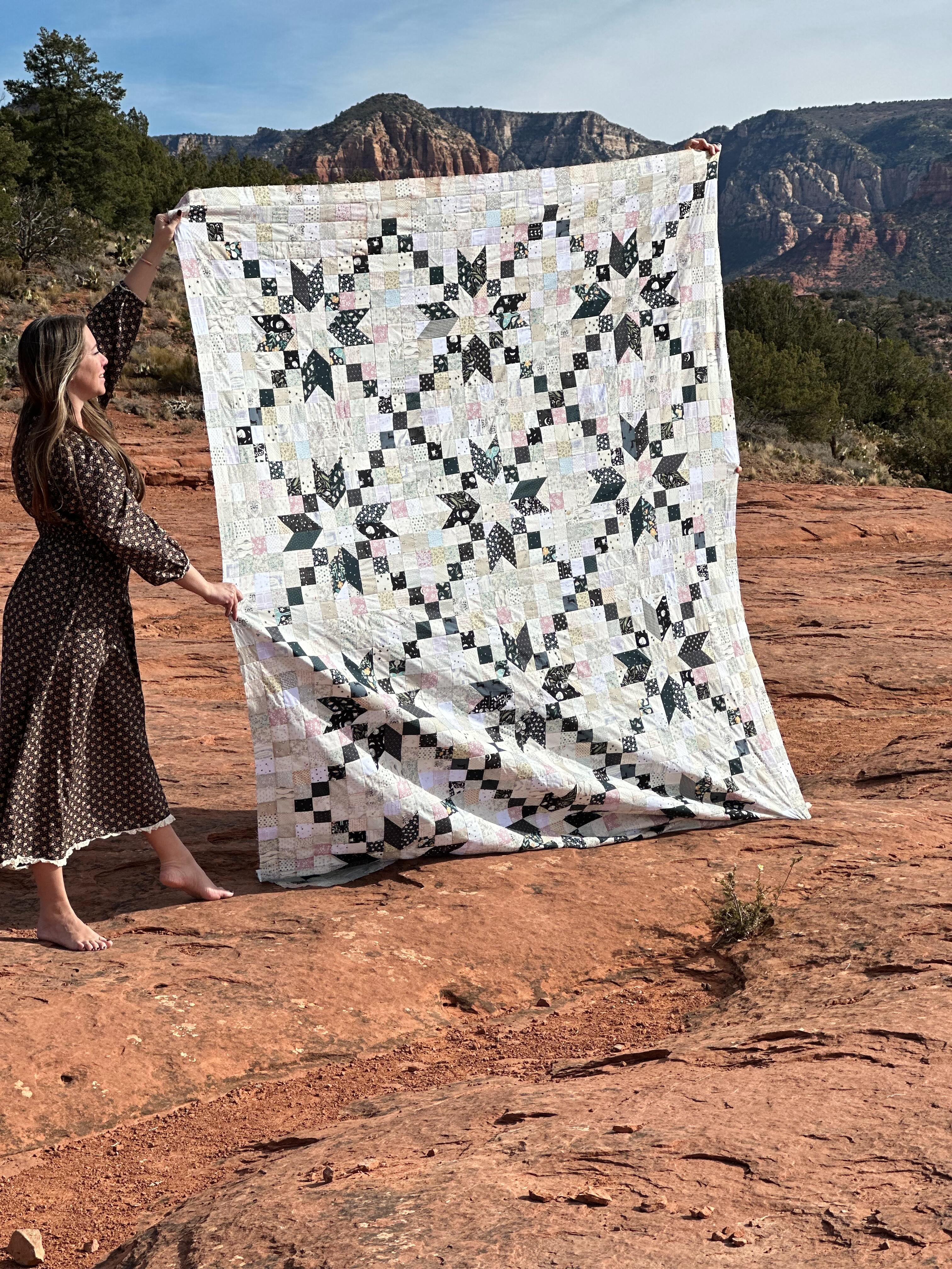 A woman in a brown polka-dot dress holds up a large quilted patchwork blanket with a black and white geometric star pattern, set against a red rock desert landscape with mountains in the background.