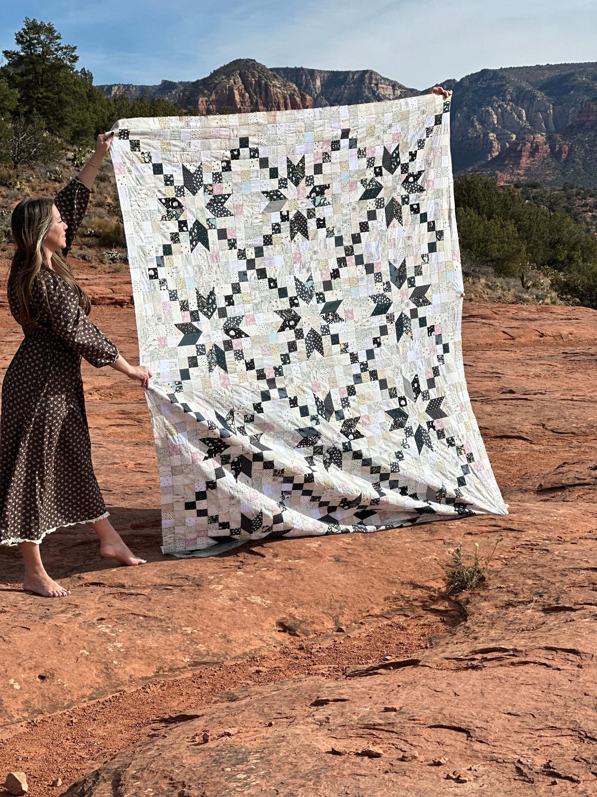 A woman in a brown polka-dot dress holds up a large quilted patchwork blanket with a black and white geometric star pattern, set against a red rock desert landscape with mountains in the background.