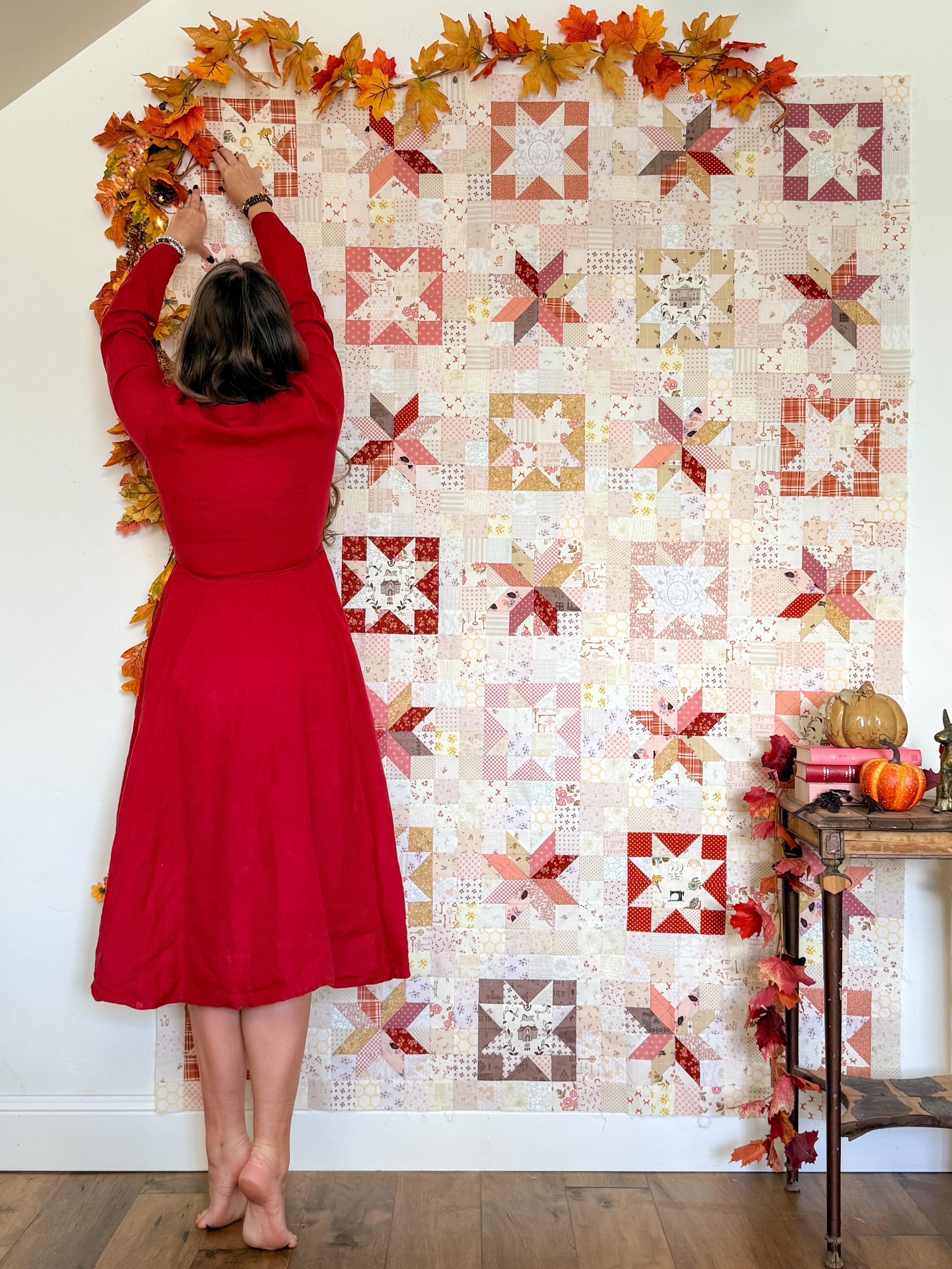 A woman in a red dress hangs autumn leaf garlands along the top edge of a large quilted wall hanging. The quilt features a patchwork design with star and geometric blocks in warm fall tones including red, orange, beige, and brown on a white background.