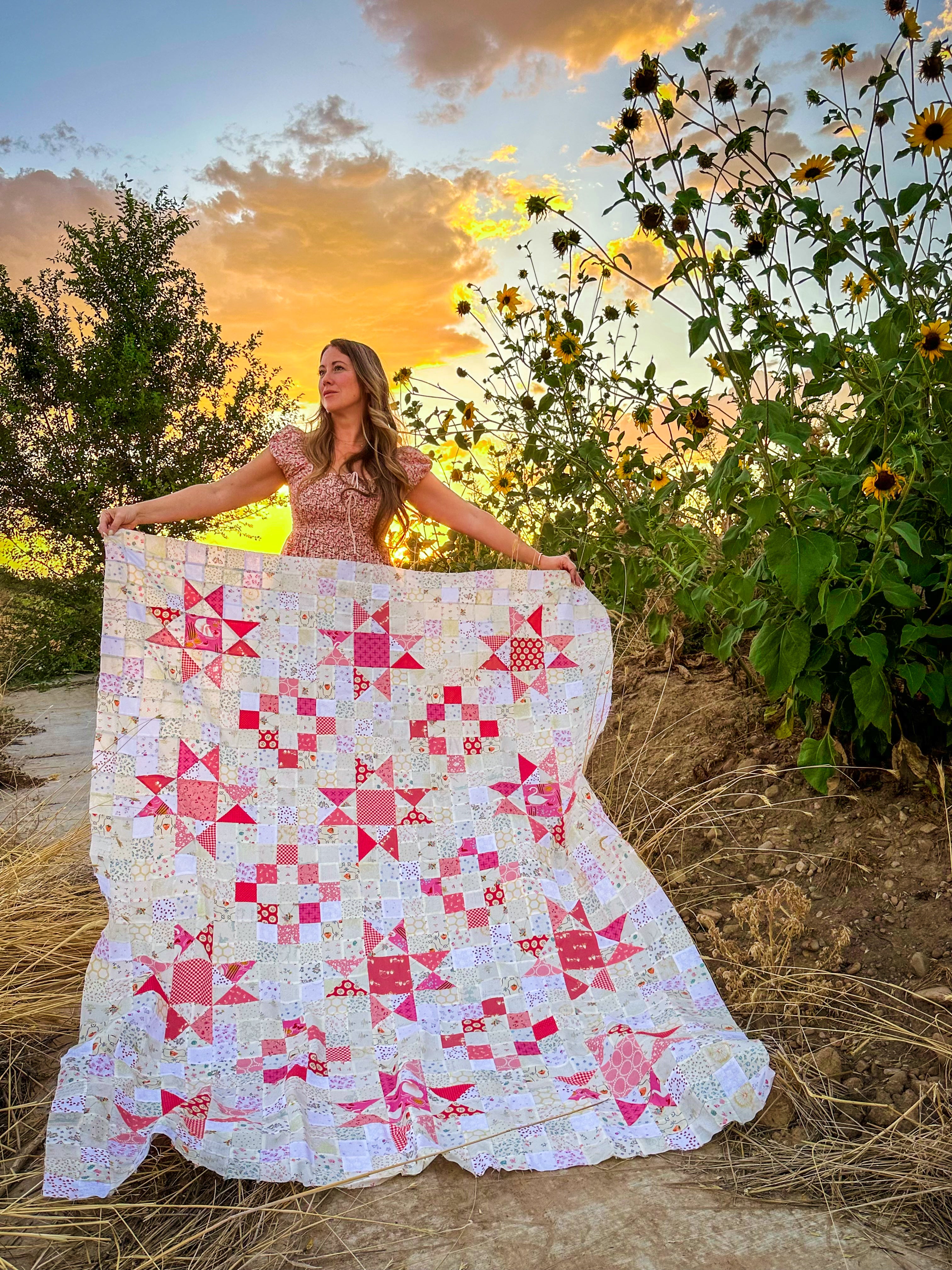 A woman outdoors at sunset holds up a large quilted blanket featuring a patchwork design of pink, red, and white star and geometric blocks, with sunflowers and trees in the background.